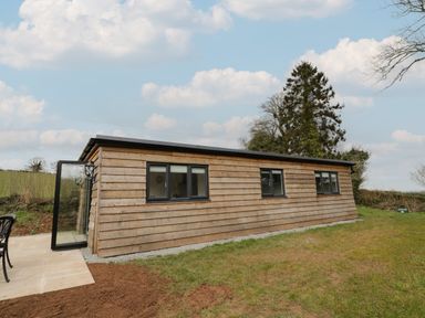 An exterior view of a wooden cabin with multiple windows and an open glass door on a grassy lawn at Goldthorne Lodge in Clee Hill
