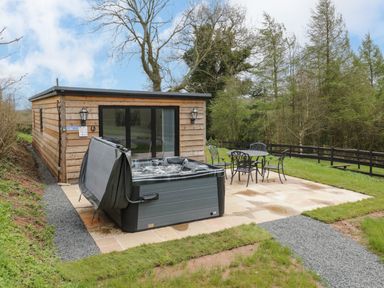 An outdoor patio with a hot tub and a metal table with four chairs next to a wooden cabin at Goldthorne Lodge in Clee Hill