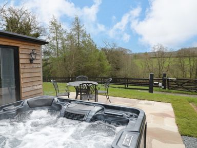 An outdoor patio with a hot tub and metal table and chairs near greenery at Goldthorne Lodge in Clee Hill
