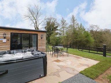 An outdoor patio area with a hot tub and metal table with four chairs next to a wooden building at Goldthorne Lodge in Clee Hill