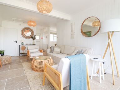 A living room with beige sofas wooden chairs rattan coffee table round mirrors and a floor lamp at Polwen in Harlyn Bay Padstow