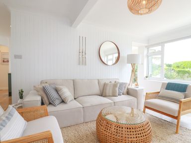 A living room with a beige sofa striped cushions rattan coffee table armchairs a floor lamp and a round mirror at Polwen in Harlyn Bay Padstow