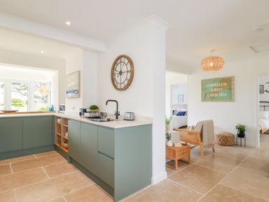 A kitchen with green cabinets and a wall clock adjacent to a sitting area with wooden furniture and a framed sign at Polwen in Harlyn Bay Padstow