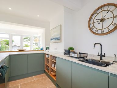 A kitchen with green cabinets a sink a large wall clock and shelves with glassware at Polwen in Harlyn Bay Padstow