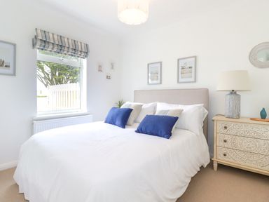 A bedroom with a bed with blue and white pillows a bedside table with a lamp and a window with striped blinds at Polwen in Harlyn Bay Padstow