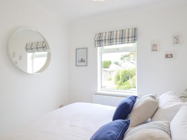A bedroom with a bed with blue and beige pillows a round mirror and a window with striped blinds at Polwen in Harlyn Bay Padstow