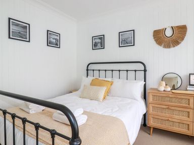 A bedroom with a black metal bed frame white bedding and a wooden dresser with decorative items and framed pictures on the walls at Polwen in Harlyn Bay Padstow
