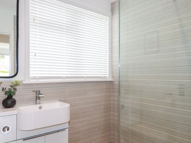 A bathroom with a white sink cabinet a window with white blinds and a glass shower wall at Polwen in Harlyn Bay Padstow