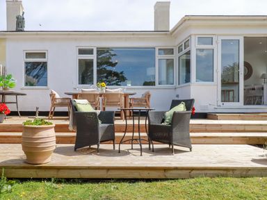 A wooden deck with two black wicker chairs and a table in front of a white house with large windows and an open door at Polwen Harlyn Bay Padstow