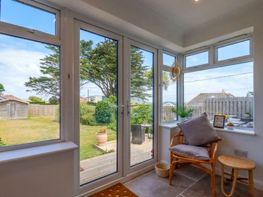 A corner room with large windows and glass doors leading to a garden with a wooden chair and cushions inside at Polwen in Harlyn Bay Padstow