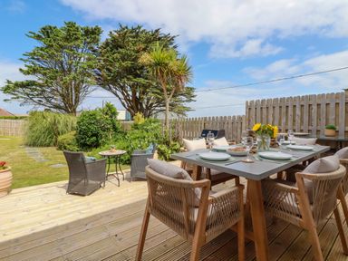 An outdoor dining area with a table set for six on a wooden deck surrounded by garden plants at Polwen in Harlyn Bay Padstow