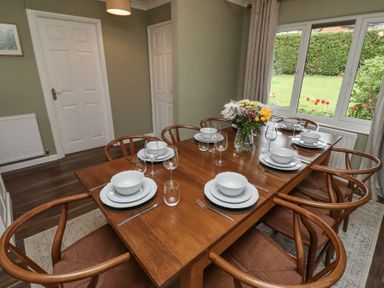 A dining room with a wooden table set with plates bowls glasses and cutlery six wooden chairs and a window with a view of a garden at Sparrow House in Alnwick