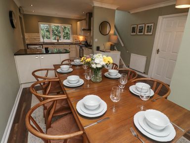 A dining area with a wooden table set with white dishes and glassware and a kitchen with counters and a window at Sparrow House in Alnwick