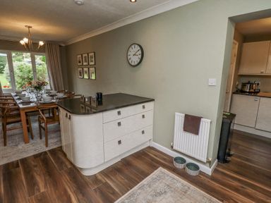 A dining area with a wooden table set with dishes and glasses next to a kitchen island with drawers and a wall clock at Sparrow House in Alnwick