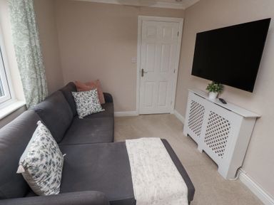 A living room with a dark gray sofa and patterned cushions a white radiator cover with a potted plant and a wall mounted television at Sparrow House in Alnwick