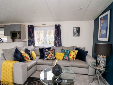 A living room with a grey corner sofa with colorful cushions a glass coffee table and side table with a lamp at The Hideout in Gilfachrheda near Llanarth