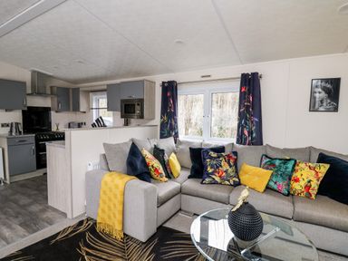 A living room with a gray sofa decorated with colorful pillows and a yellow throw next to a glass coffee table and a kitchen area at The Hideout in Gilfachrheda near Llanarth