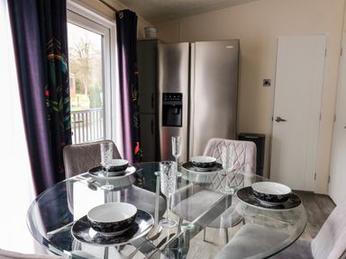 A dining area with a glass table set for four with bowls plates and glasses next to a window with dark curtains and a double stainless steel refrigerator at The Hideout in Gilfachrheda near Llanarth