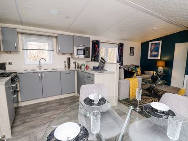 A kitchen and living area with grey cabinets a glass dining table set and a sofa with colorful cushions at The Hideout in Gilfachrheda near Llanarth