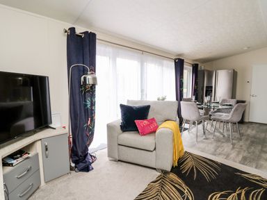 A living area with a gray armchair, TV on a stand, dark curtains, glass dining table with chairs, and a refrigerator in the background at The Hideout in Gilfachrheda near Llanarth
