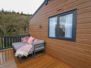 An outdoor wooden deck with a gray sectional sofa with pink pillows and a blanket next to a house with a window at The Hideout in Gilfachrheda near Llanarth