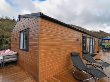 A wooden deck with two black lounge chairs a sofa with cushions and a dining table with an umbrella at The Hideout in Gilfachrheda near Llanarth