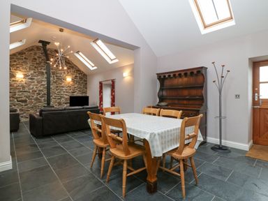 A dining area with wooden chairs and table near a living room with stone wall and black sofas at The Old Dairy in Sarn Bach near Abersoch