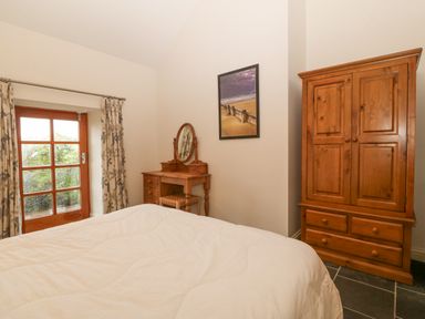 A bedroom with a wooden wardrobe a dressing table a bed and a window with floral curtains at The Old Dairy in Sarn Bach near Abersoch