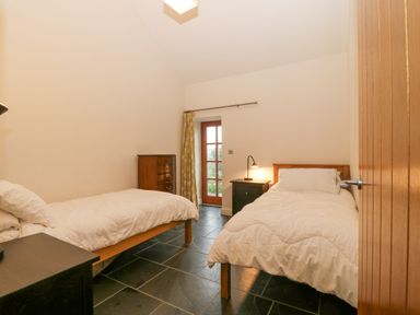 A bedroom with two single beds a chest of drawers a bedside table with a lamp and a door with curtains at The Old Dairy Sarn Bach near Abersoch