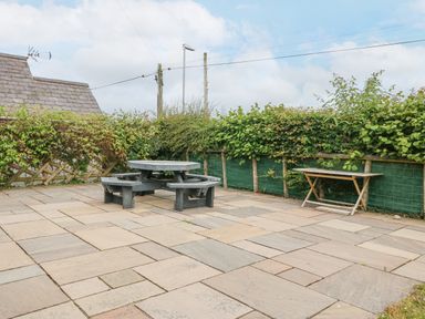 An outdoor patio area with a round picnic table and benches and a folding table against a hedge at The Old Dairy in Sarn Bach near Abersoch