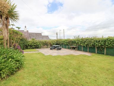 A garden with grass a patio with a round picnic table and benches plants and a trees with a hedge in the background at The Old Dairy in Sarn Bach near Abersoch