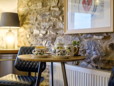 A round wooden table with floral teacups teapot and cake near a stone wall and black quilted chairs at Sunny Brae in Lindale