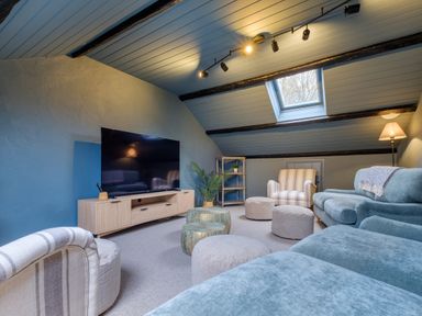 An attic living room with a large television on a wooden stand a blue sofa striped armchairs round poufs side tables and a skylight window at Sunny Brae in Lindale