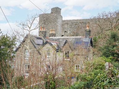 Stone house with chimneys and solar panels surrounded by trees with a large stone castle wall in the background at Hafod Y Bryn in Harlech