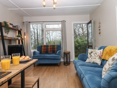 A living room with blue sofas a wooden table with orange juice and bread and a shelf with books at 2 The Glade Kilkhampton