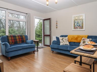 A living room with two blue sofas a wooden dining table with croissants and large windows showing trees at 2 The Glade in Kilkhampton