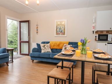 A living room with blue sofas and a wooden dining table set with croissants and glasses of orange juice at 2 The Glade in Kilkhampton