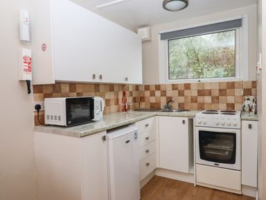 A kitchen with white cabinets a microwave a small refrigerator an oven a sink and a window with a view of trees at 2 The Glade in Kilkhampton