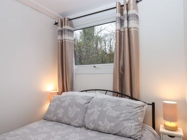 A bedroom with a bed featuring grey floral bedding two bedside lamps and a window with beige curtains at 2 The Glade in Kilkhampton