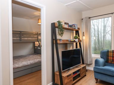 A room with bunk beds visible through a doorway a shelving unit with a television and books and a blue armchair by a window at 2 The Glade in Kilkhampton