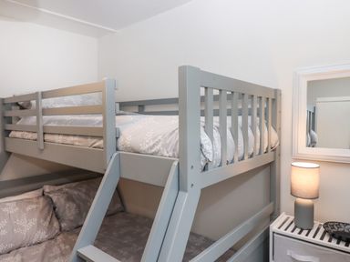 A bedroom with a gray bunk bed a lamp on a white side table and a mirror on the wall at 2 The Glade in Kilkhampton