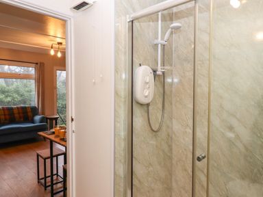A shower area with a wall-mounted shower unit and sliding glass door next to a living room with a sofa and a table at 2 The Glade in Kilkhampton