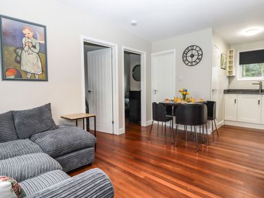 A living area with a gray sofa a small wooden side table a round dining table with four black chairs a wall clock and a kitchen area with white cabinets at 15 The Park in Kilkhampton