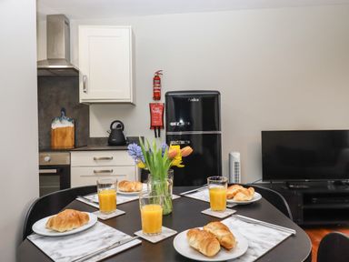 A kitchen and dining area with a black refrigerator a kettle on the countertop a table set with plates of croissants glasses of orange juice and a vase of flowers at 15 The Park in Kilkhampton