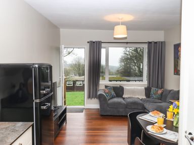 A living room with a grey corner sofa a black refrigerator a dining table with croissants and glasses of orange juice a door leading outside and windows with grey curtains at 15 The Park in Kilkhampton