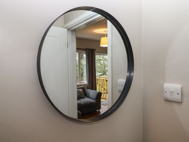 A round mirror on a wall reflecting a room with a grey sofa a window with curtains and a hanging light at 15 The Park in Kilkhampton