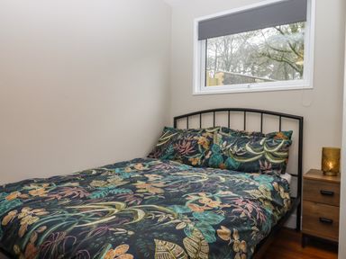 A bedroom with a bed featuring floral bedding a wooden nightstand with a lamp and a window showing trees at 15 The Park in Kilkhampton