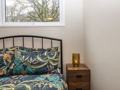 A bedroom with a bed with leaf patterned bedding and a wooden nightstand with a decorative lamp at 15 The Park in Kilkhampton
