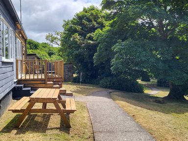 A wooden picnic table on lawn beside a paved path and a house with a deck and chairs surrounded by trees at 15 The Park in Kilkhampton