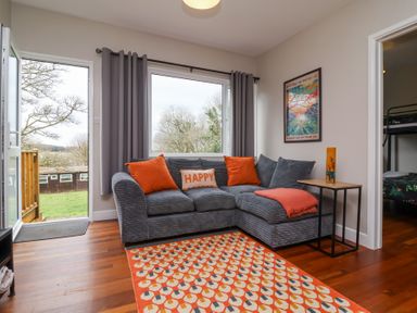 A living room with a gray sectional sofa with orange cushions and a patterned orange rug at 15 The Park in Kilkhampton